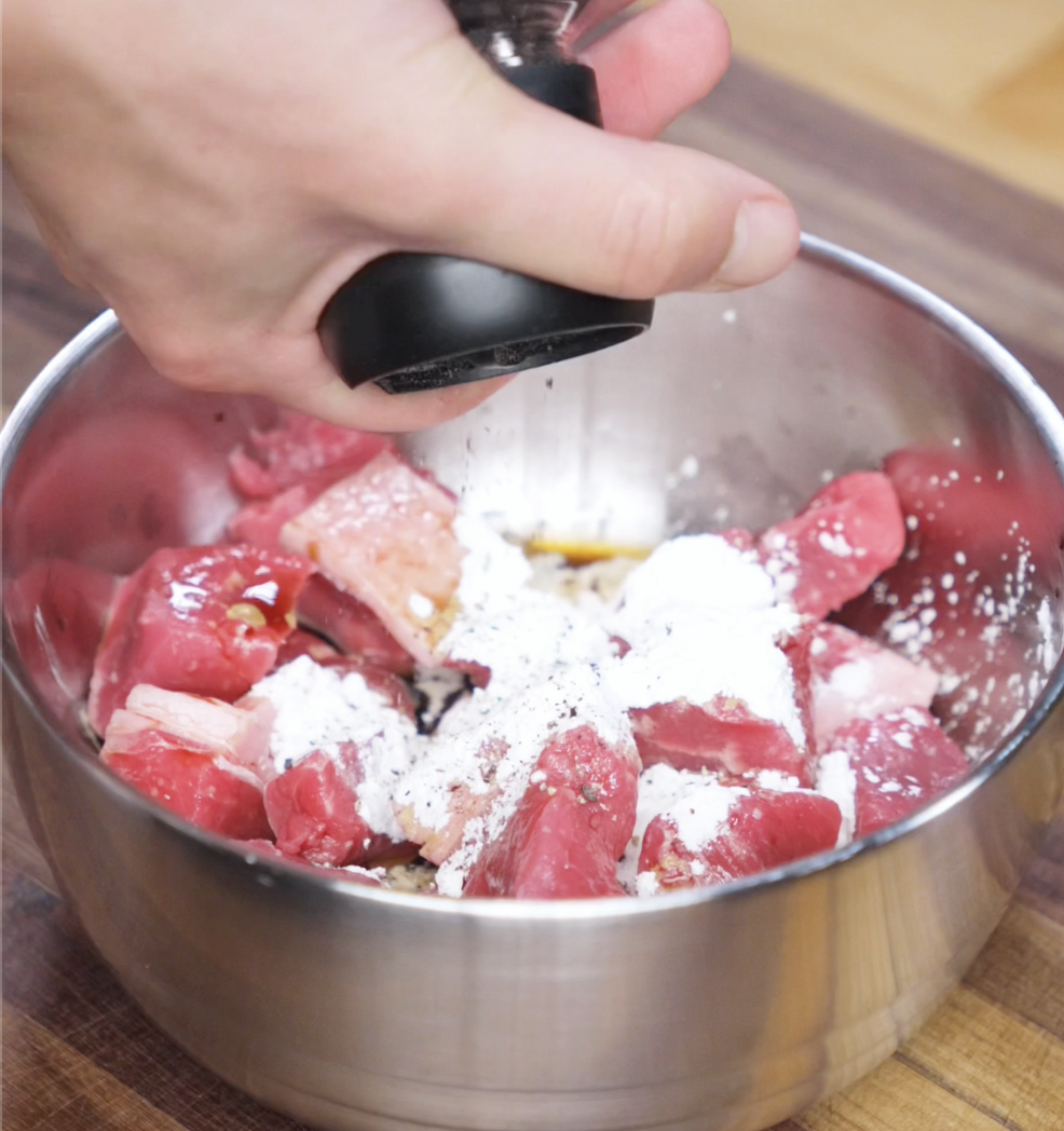 A bowl of raw beef cubes being marinated with soy sauce, shaoxing wine, cornstarch, and black pepper. A hand sprinkles fresh black pepper over the top, readying the beef for a bold stir-fry.