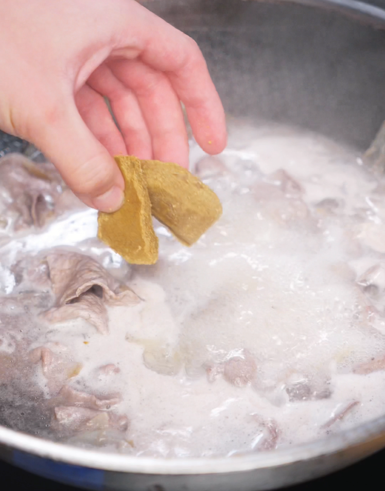 A hand drops two blocks of golden Japanese curry roux into a pot of simmering beef and water. The curry base begins to melt and thicken into a rich, flavorful sauce.