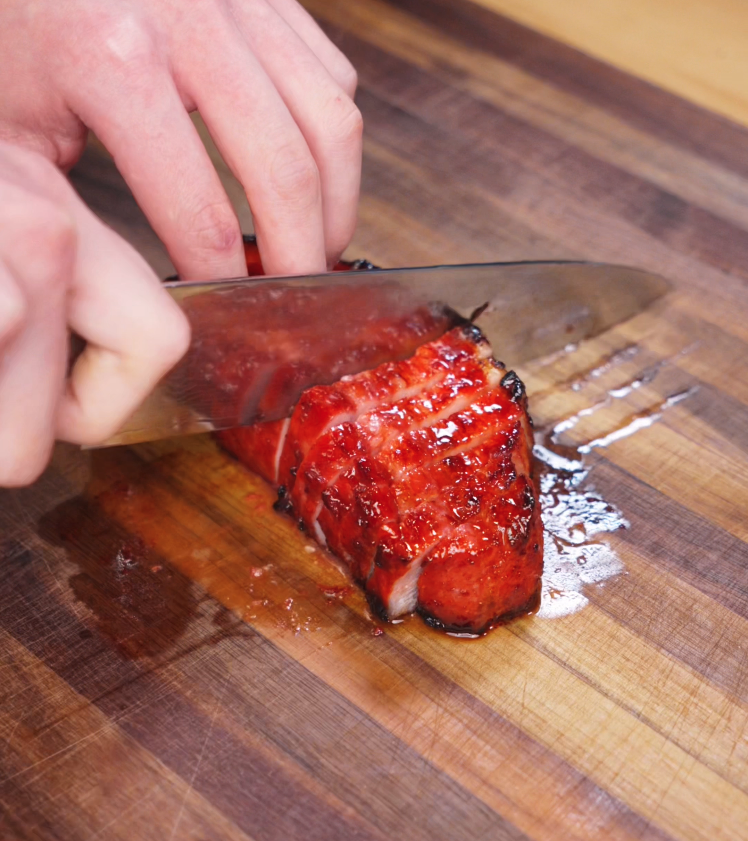 A knife glides through juicy pork belly, revealing the caramelized outer layer and tender interior. The slices are evenly cut and ready to be served over rice.