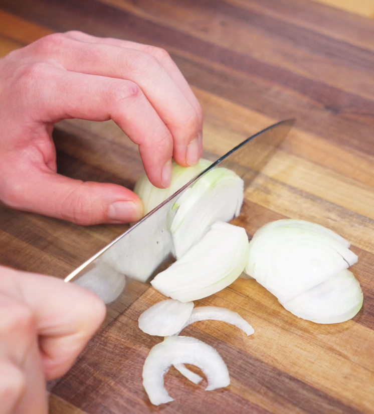 A hand holds a knife while slicing a white onion into thin half-moons on a wooden cutting board. These onions will add sweetness and depth to the savory Japanese curry broth.