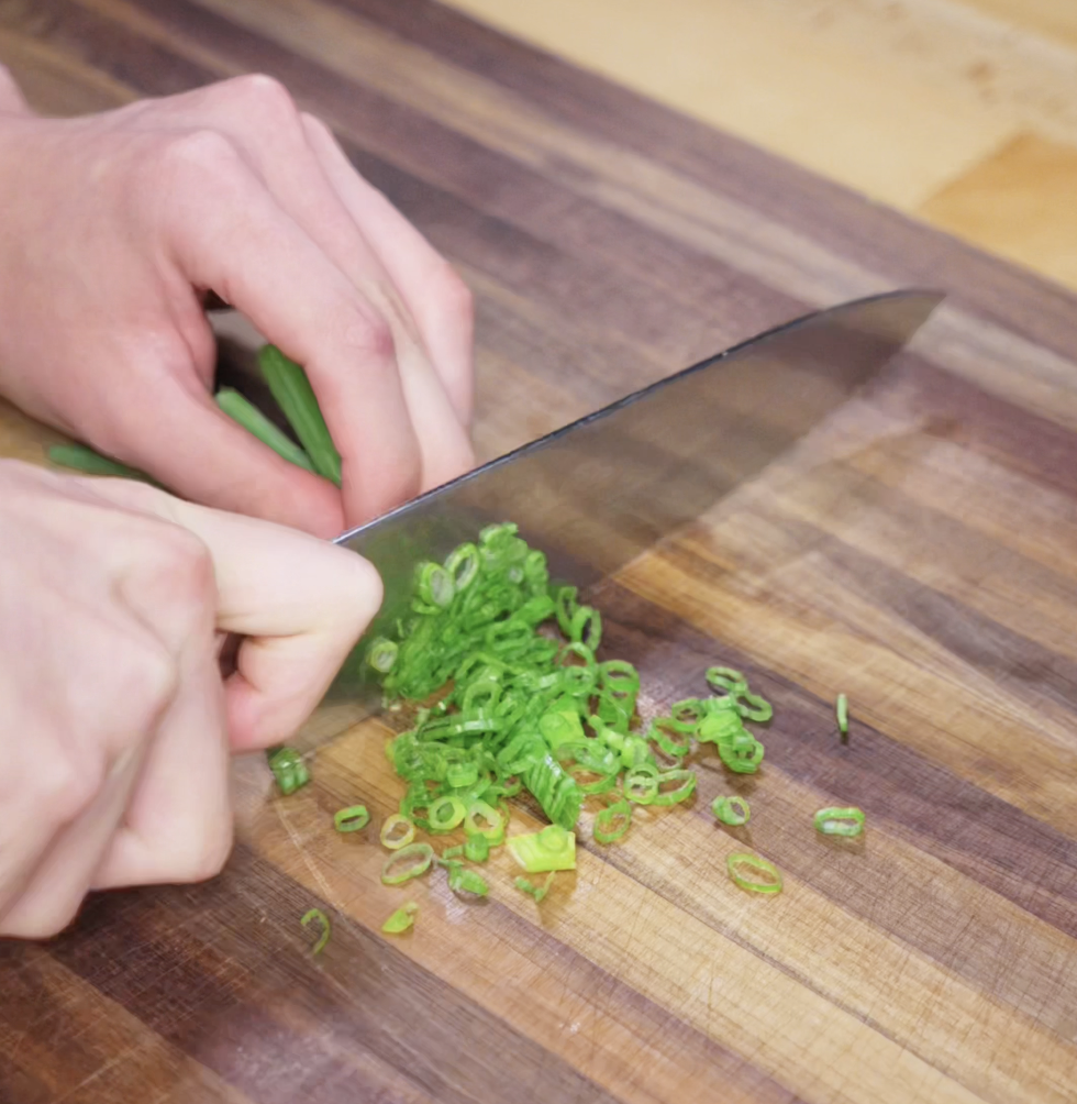 A hand slices bright green scallions into thin rings on a wooden cutting board. The scallions will add fresh flavor and color contrast to the final fried rice dish.