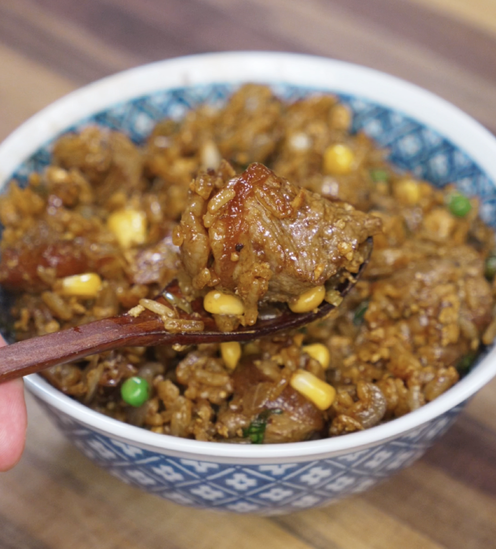 A wooden spoon lifts a tender, glazed cube of beef along with saucy rice, corn, and peas. The beef is caramelized and juicy, coated in bold black pepper sauce, highlighting the dish’s hearty texture.