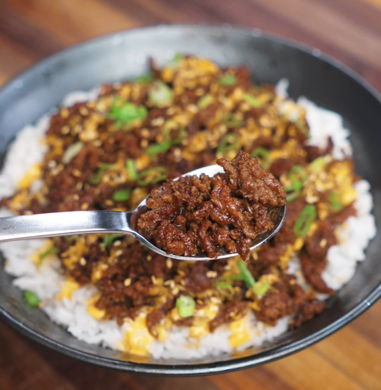 A close-up of a spoon lifting a bite of caramelized Japanese BBQ ground beef above a bowl of fluffy white rice. The beef is rich in color, glossy with sauce, and speckled with sesame seeds and scallions, showcasing its savory, umami-packed coating.