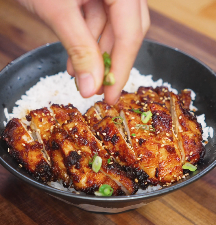 A hand sprinkles chopped scallions over a finished rice bowl topped with sliced peanut glazed chicken. The glistening chicken is adorned with sesame seeds and sits atop fluffy rice, ready to serve.