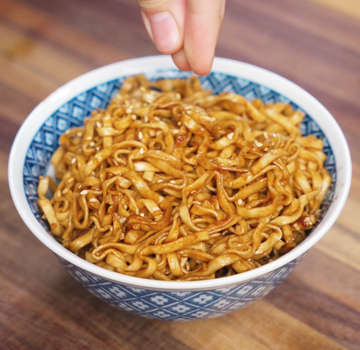 A hand sprinkles toasted sesame seeds over a finished bowl of garlic butter noodles, adding a nutty crunch and final garnish.