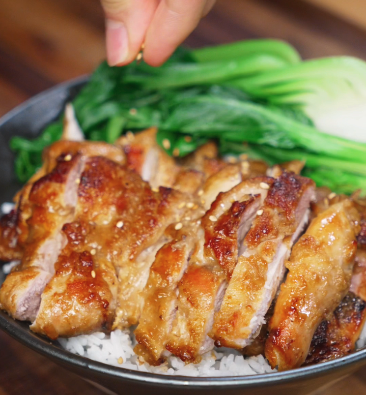 A hand sprinkles sesame seeds over the lemon pepper chicken and rice bowl. The toasted seeds add a nutty flavor and visual contrast to the golden chicken and greens.