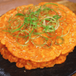A close-up of golden, crispy kimchi pancakes stacked on a black plate. The pancakes are garnished with finely sliced scallions and toasted sesame seeds, highlighting their crispy texture and vibrant color.