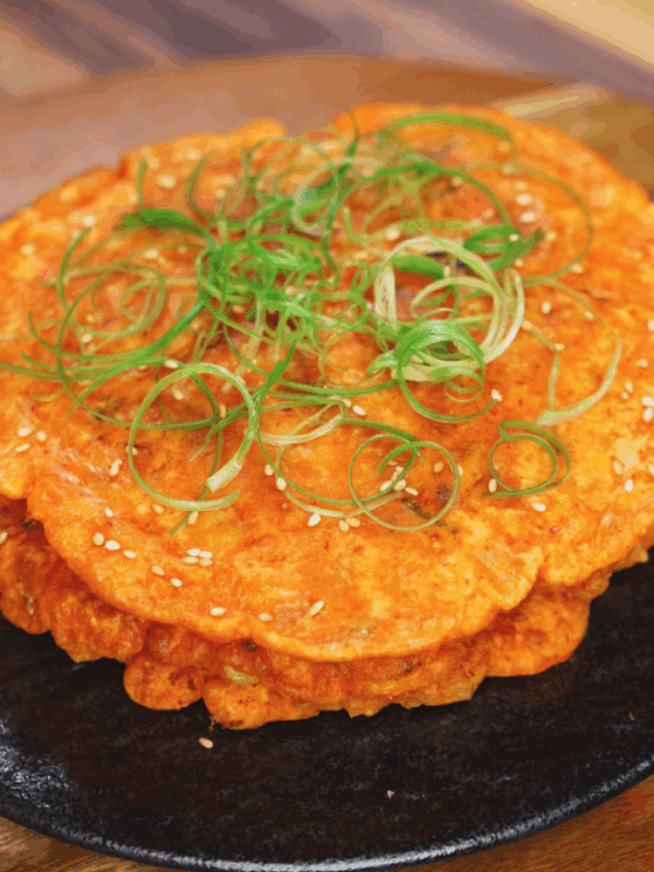 A close-up of golden, crispy kimchi pancakes stacked on a black plate. The pancakes are garnished with finely sliced scallions and toasted sesame seeds, highlighting their crispy texture and vibrant color.