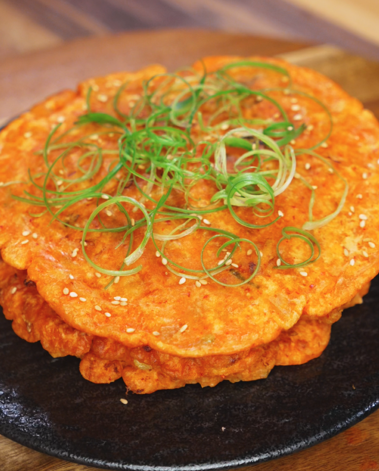 A close-up of golden, crispy kimchi pancakes stacked on a black plate. The pancakes are garnished with finely sliced scallions and toasted sesame seeds, highlighting their crispy texture and vibrant color.
