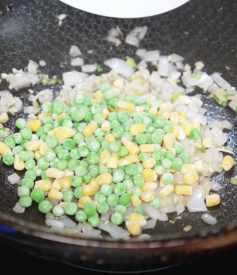 A mix of diced onion, garlic, peas, and corn cooks in a hot pan. The vegetables begin to soften and release their aroma, forming the savory base of the fried rice.