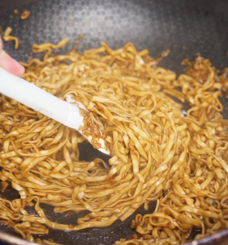 A spatula stirs cooked noodles in a skillet, evenly coating them in glossy garlic butter sauce infused with soy, oyster sauce, and aromatics.