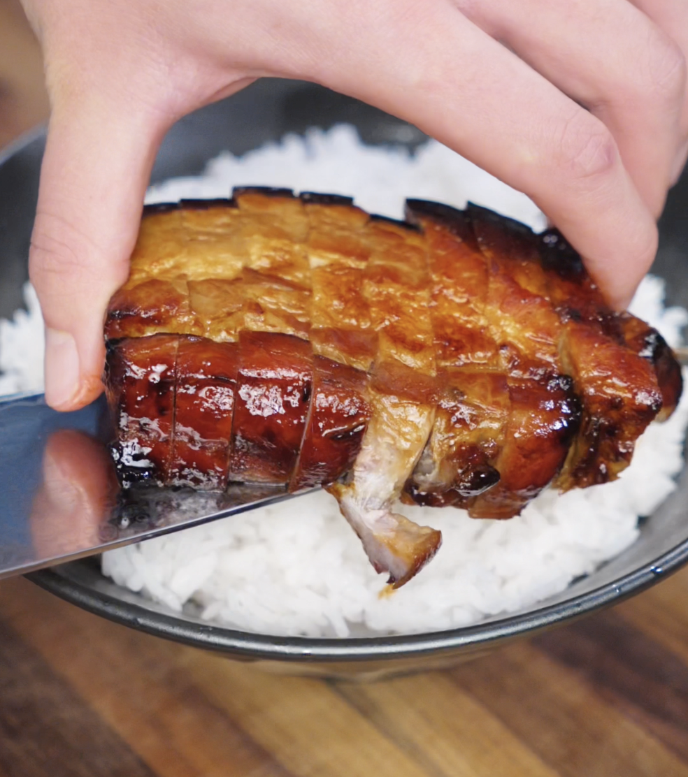 A hand carefully places a sliced slab of glazed pork belly onto a bowl of white rice using a knife. The glossy exterior contrasts with the fluffy rice base underneath.