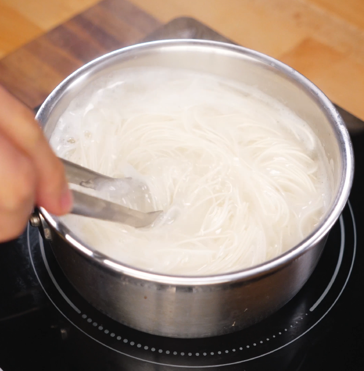 Cooking somyeon (thin wheat noodles) in boiling water inside a stainless steel pot, stirred with tongs to prevent sticking.