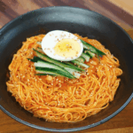 Overhead view of a black bowl filled with spicy cold noodles, topped with cucumber slices, chopped kimchi, and half a soft-boiled egg sprinkled with sesame seeds.