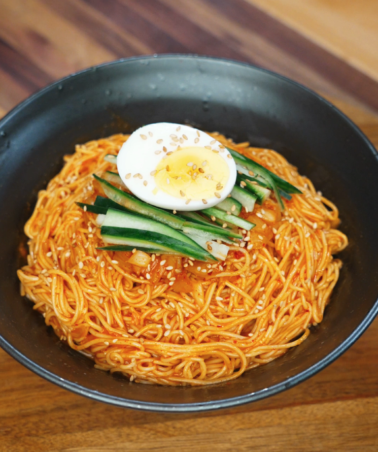 Overhead view of a black bowl filled with spicy cold noodles, topped with cucumber slices, chopped kimchi, and half a soft-boiled egg sprinkled with sesame seeds.