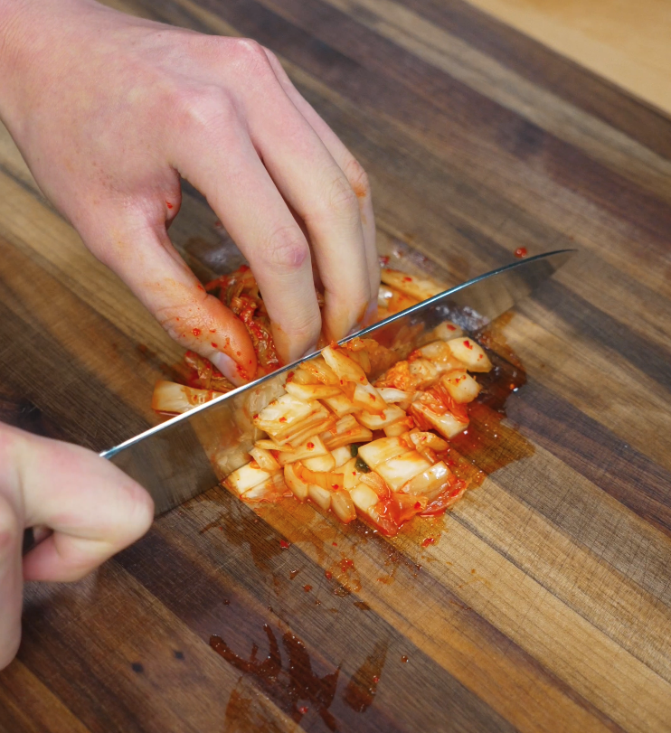 Hand chopping fresh kimchi on a wooden cutting board, showcasing the juicy, fermented cabbage pieces and bright red chili marinade.