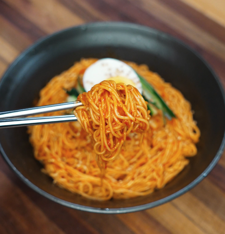 Close-up of chopsticks lifting a tangle of bibim guksu noodles coated in bright red spicy sauce, highlighting the texture and vibrant color of the cold Korean dish.