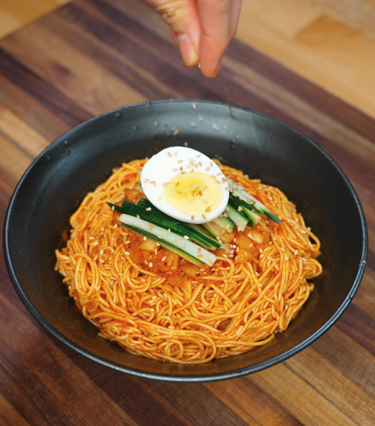 Sprinkling sesame seeds over a completed bowl of bibim guksu, garnished with julienned cucumbers, chopped kimchi, and half a boiled egg.