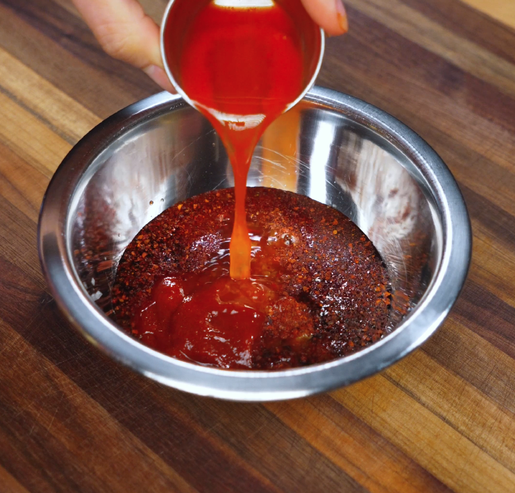 Pouring red gochujang-based sauce into a metal mixing bowl filled with Korean chili flakes and other seasoning ingredients for bibim guksu.