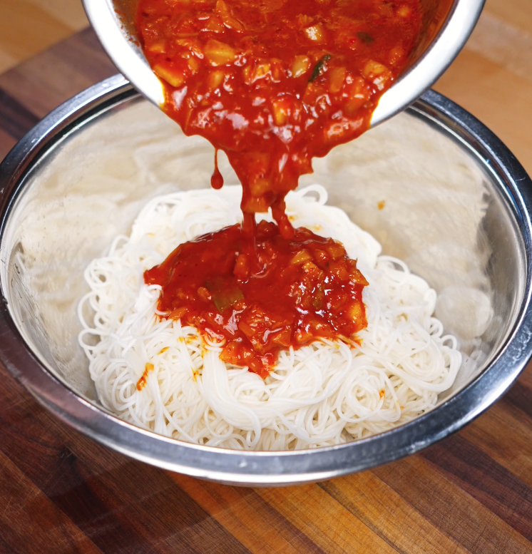 Pouring spicy kimchi gochujang sauce over a bowl of freshly boiled and rinsed cold noodles, preparing to mix the bibim guksu.