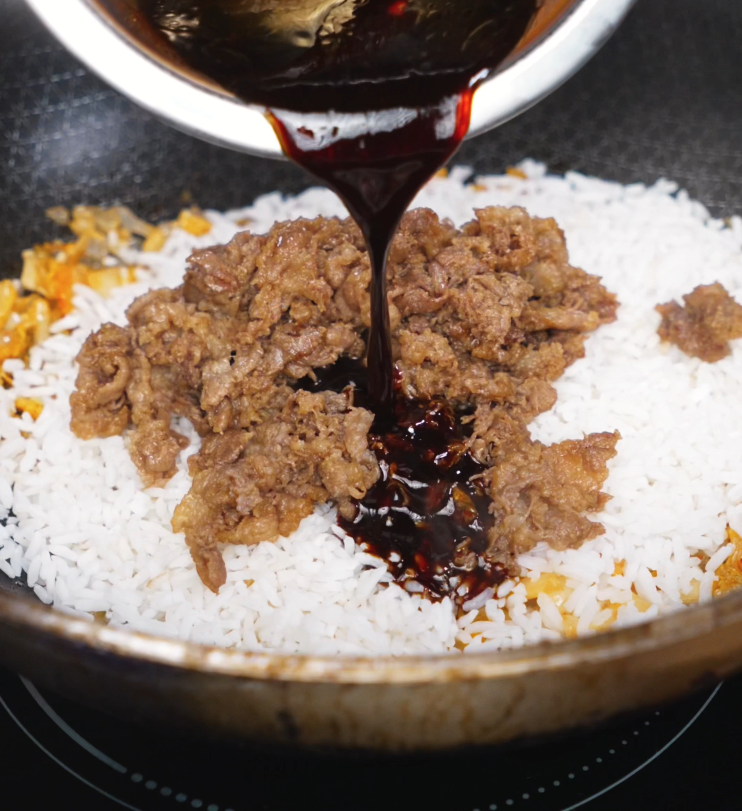 A rich, dark sauce is poured over white rice and cooked beef in a skillet, bringing the final components together for stir-frying.