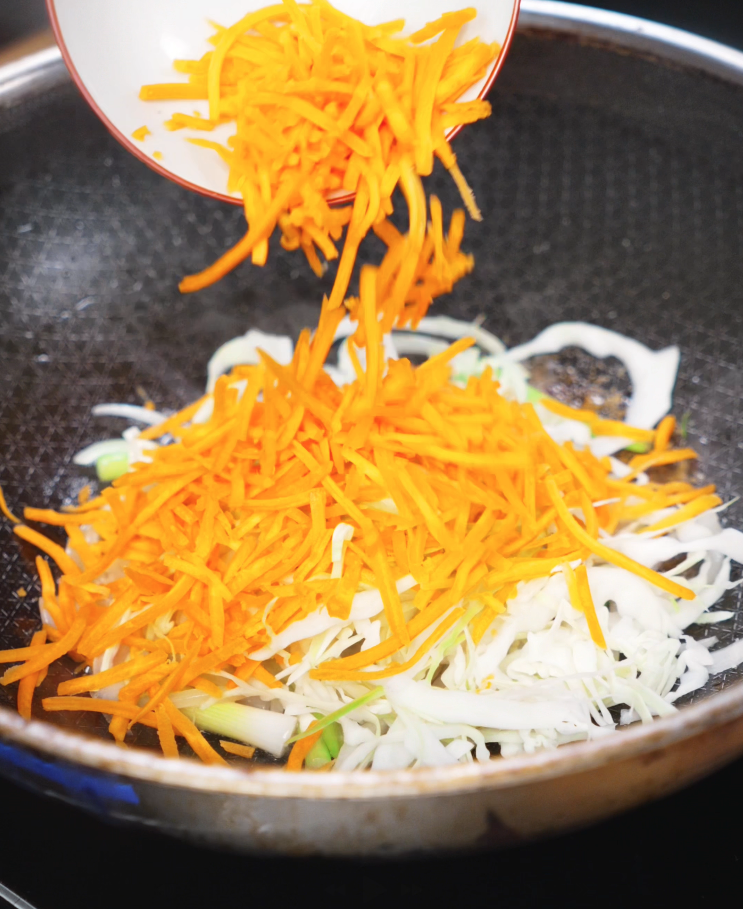 Shredded carrots and sliced cabbage are added to a hot pan of onions and scallions to create a colorful veggie mix for the stir-fry.