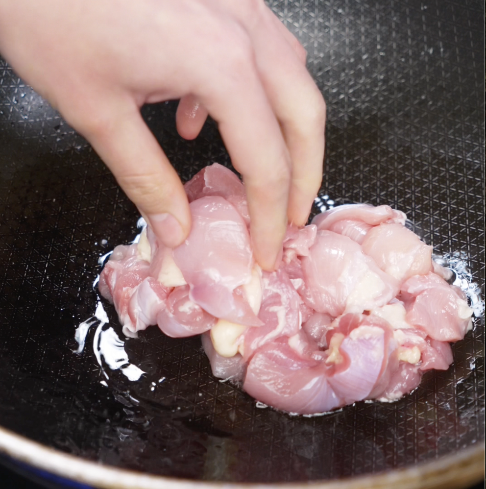 Raw chicken chunks are placed into a lightly oiled pan, beginning the cooking process for the garlic chicken fried rice.