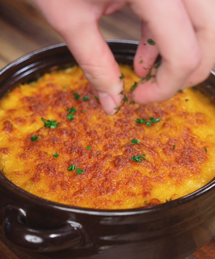 A hand sprinkles chopped fresh parsley over a bubbling, golden-brown baked mac and cheese topped with a perfectly crisp cheese crust.