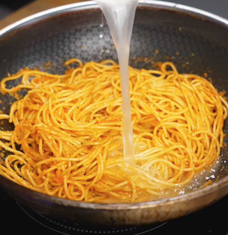 A stream of reserved pasta water is poured into the gochujang-coated noodles, helping to emulsify the sauce to a silky consistency.