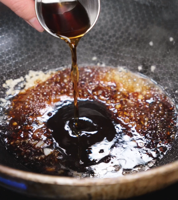A dark soy-based sauce is poured into the pan of garlic, combining into a rich, savory glaze.