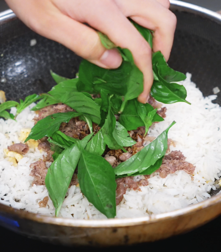 A handful of fresh Thai basil leaves is added to the hot pan of rice and beef, releasing a burst of fragrance as they wilt into the dish.