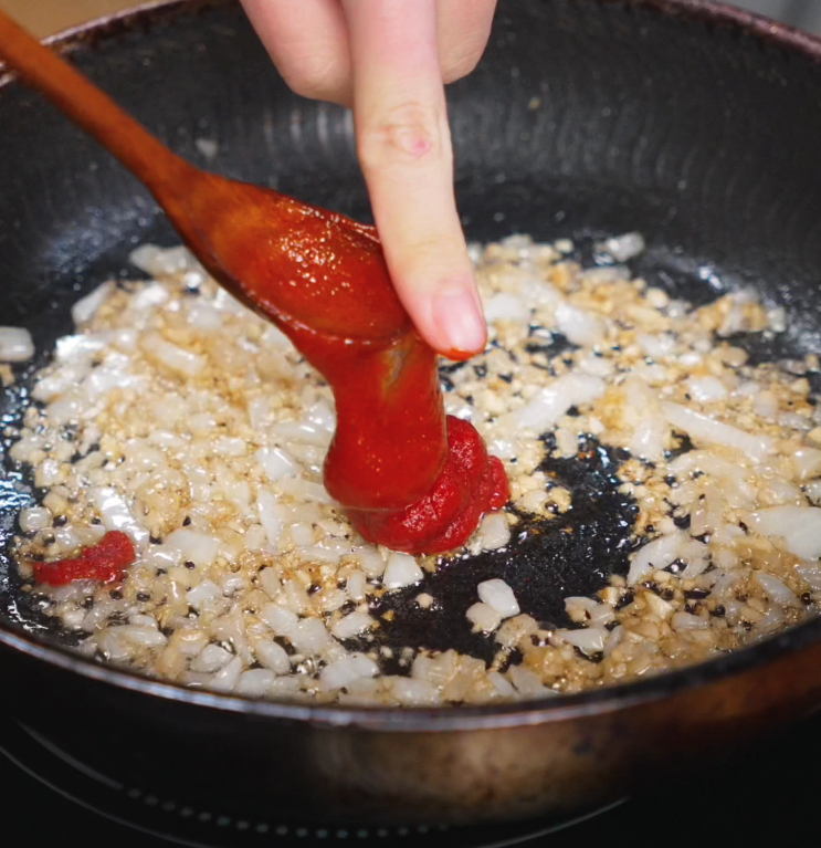 Tomato paste is pushed off a wooden spoon into a skillet of sautéed onions and garlic, beginning the base of a rich, spicy sauce.