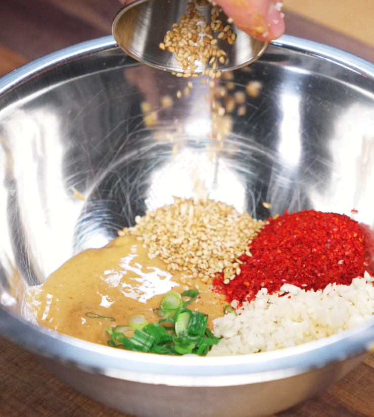 A metal bowl holds peanut butter, chopped scallions, chili flakes, sesame seeds, garlic, and ginger. These aromatics will be bloomed with hot oil to create a rich, nutty chili sauce.