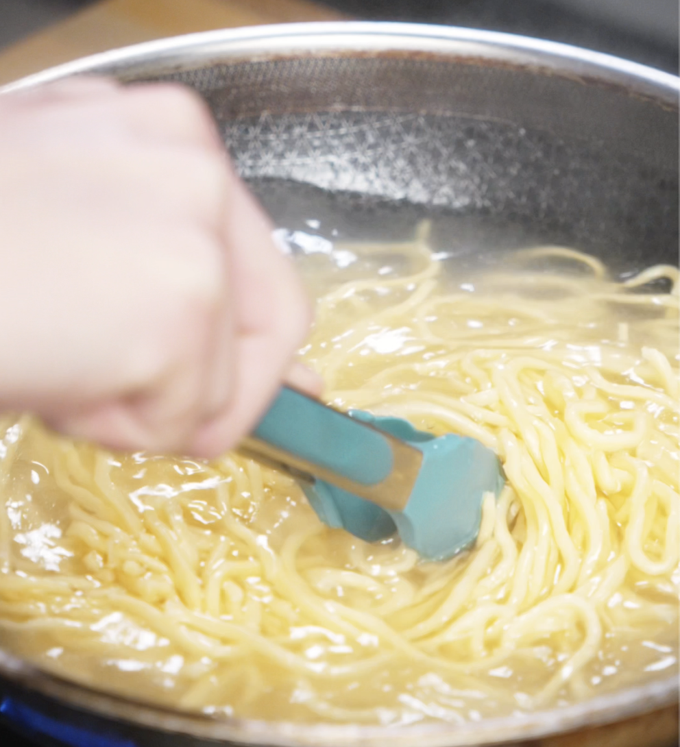 Fresh wheat noodles are boiling in a pot of water, stirred with tongs to prevent sticking. These chewy noodles form the base of the pan-fried dish.