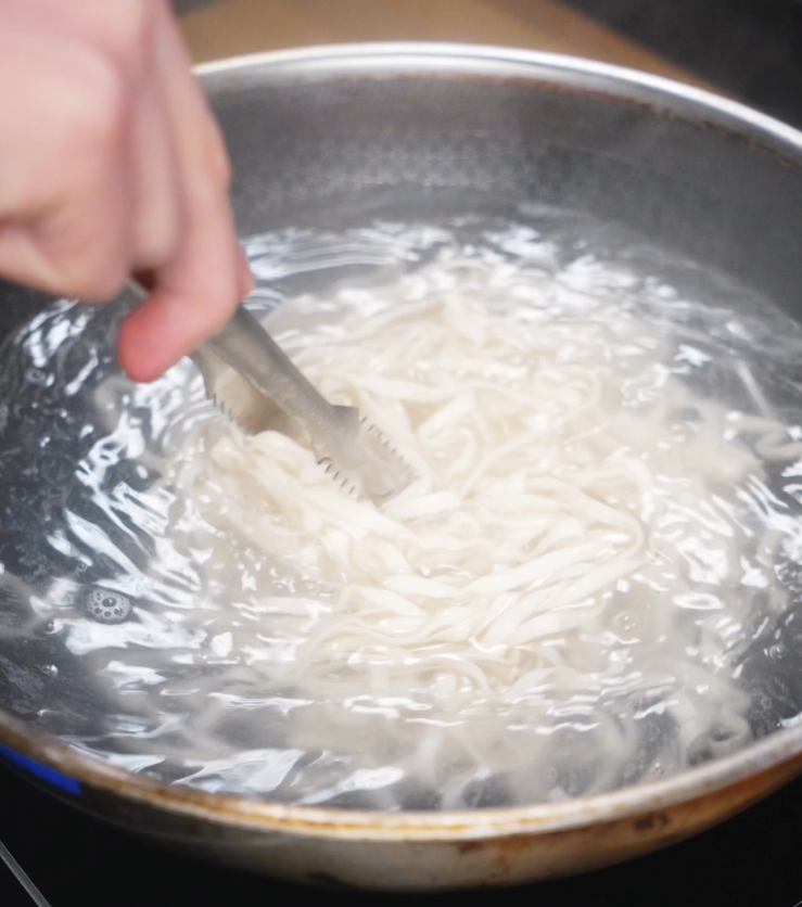 Fresh noodles are stirred in boiling water with tongs, cooking until just tender before being tossed in the sesame peanut sauce.
