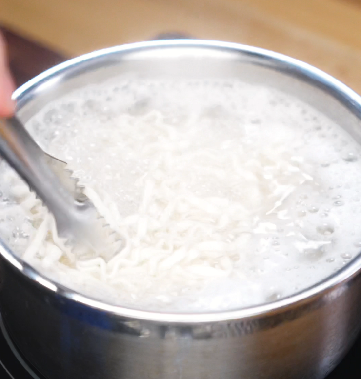 A hand uses tongs to stir noodles as they cook in a pot of boiling water. These noodles will be rinsed and tossed in the homemade spicy peanut chili oil sauce.