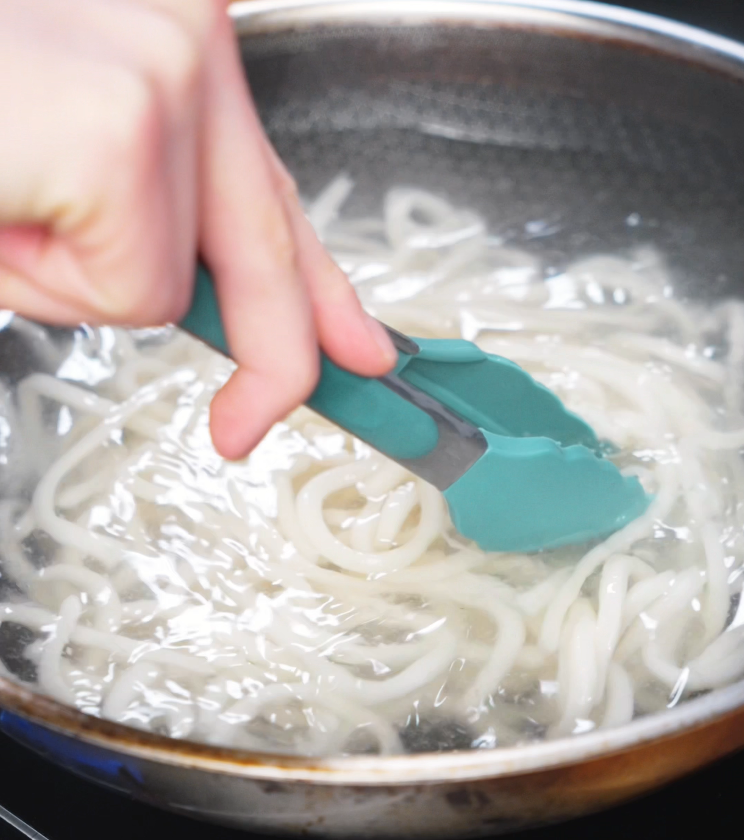 A pair of teal tongs stirs thick udon noodles in boiling water to prepare them for the chicken yaki udon stir-fry.