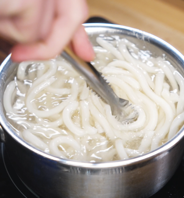 Thick udon noodles are stirred with tongs as they boil in a pot of water, being cooked just shy of al dente.