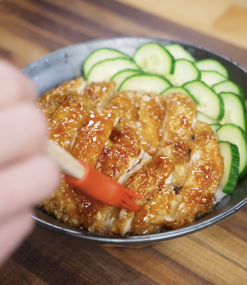 A hand uses a red brush to apply extra honey garlic glaze over sliced crispy chicken thighs, adding shine and flavor to the final rice bowl topped with cucumbers.