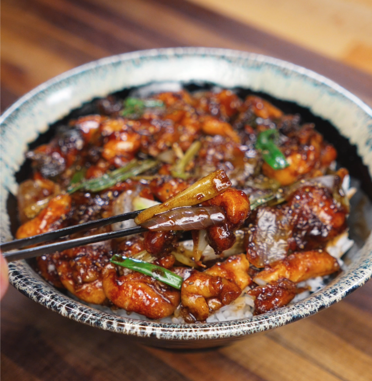 A pair of chopsticks lifts a glistening piece of black pepper chicken with onions and scallions from a bowl of rice.