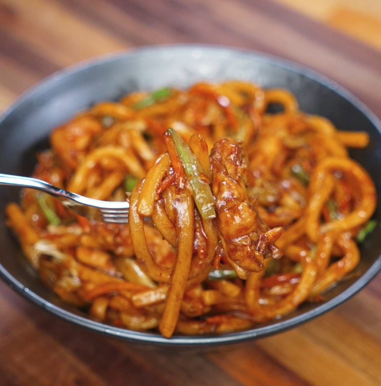 A fork lifts a saucy bite of chicken yaki udon with glistening noodles and stir-fried vegetables, showing the glossy texture of the stir-fry.