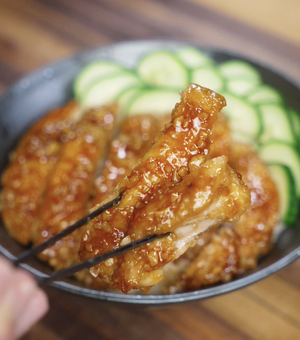 A piece of crispy chicken coated in honey garlic glaze is held up with chopsticks, highlighting the juicy interior and glossy, crunchy exterior.
