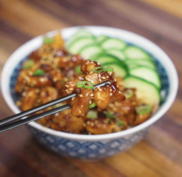 A pair of chopsticks holds up a glistening piece of garlic butter chicken garnished with scallions and sesame seeds, highlighting the caramelized sauce.