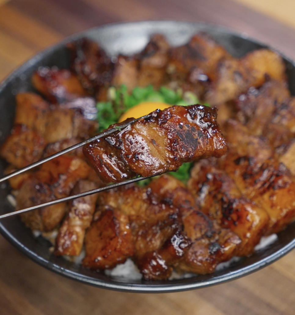 A glossy piece of caramelized pork belly is held up with chopsticks over the rice bowl, showcasing its seared edges and sticky glaze.