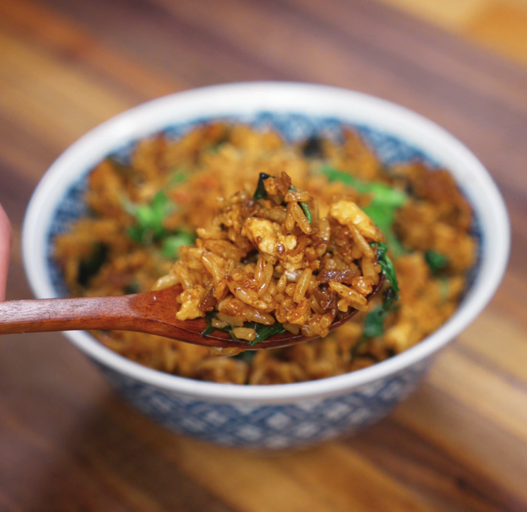 A wooden spoon lifts a bite of Thai basil fried rice showing glistening grains coated in sauce, pieces of basil, and tender beef.