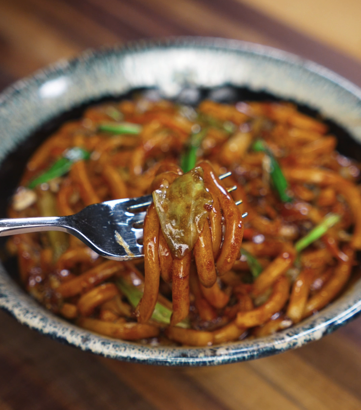 A fork lifts a twirl of thick udon noodles and a caramelized cabbage leaf, showing off the sticky teriyaki glaze clinging to each strand.