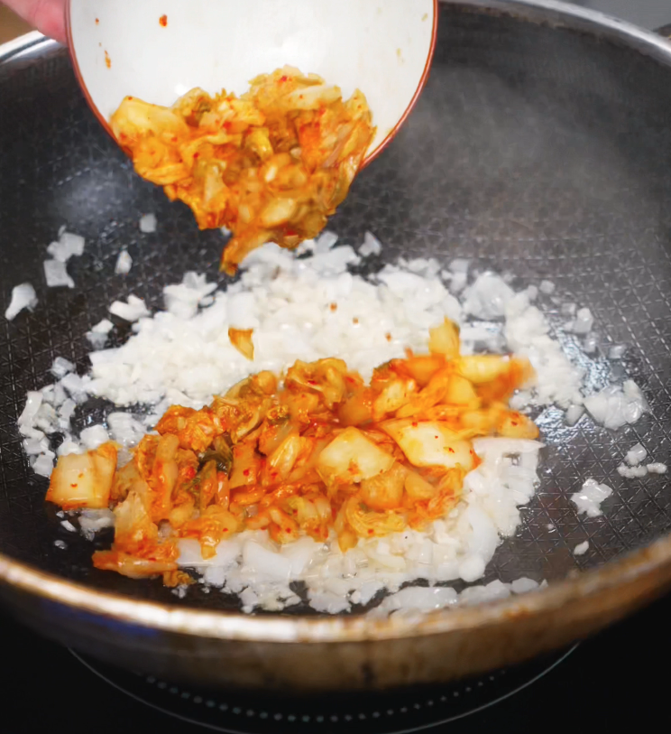 Chopped kimchi is added to sautéed onions in a hot pan, filling the kitchen with spicy, tangy aroma.