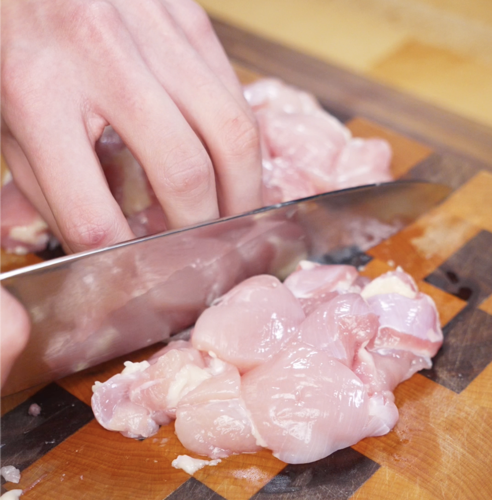 Raw boneless chicken thighs are being sliced into ½-inch chunks on a wooden cutting board, showing clean cuts and preparation for stir-frying.