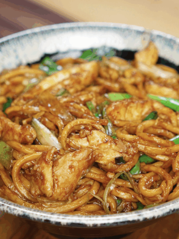A bowl is filled with chicken pan fried noodles, coated in a dark glossy sauce and mixed with scallions, onions, and cabbage. This comforting stir-fry is ready to serve.