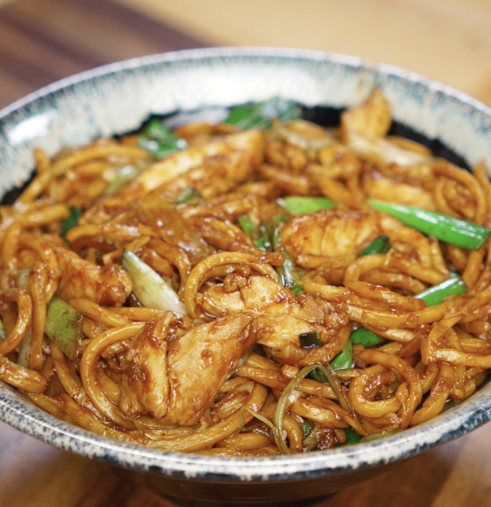A bowl is filled with chicken pan fried noodles, coated in a dark glossy sauce and mixed with scallions, onions, and cabbage. This comforting stir-fry is ready to serve.